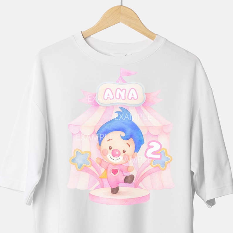 May include: White t-shirt featuring a watercolor illustration of a clown character inside a circus tent. The design includes the name "ANA" and the number "2". The clown is wearing a pink outfit and has blue hair.