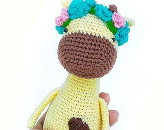 May include: A handmade crochet giraffe doll in yellow and brown, adorned with a floral crown of blue, pink, and green. The doll is held in a hand, showcasing its soft texture and detailed craftsmanship. A charming, whimsical toy.