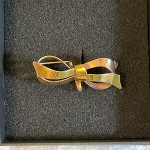 May include: A gold-toned bow brooch with a pin closure, presented in a black box. The brooch features a layered design with a polished finish, showcasing a classic bow shape. The bow's design incorporates both gold and brass tones.