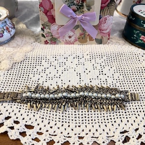 May include: A bronze-toned bracelet featuring a row of small, light-colored pearls and a series of metal spikes. The bracelet has a clasp closure and is set against a white lace doily.