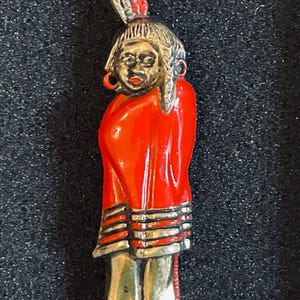 May include: A vintage brooch depicting a Native American figure. The figure is clad in a vibrant red garment with silver and white accents. The face is detailed with a silver headdress and red accents. The brooch is likely made of metal.