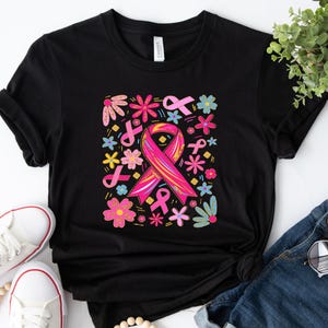 May include: Black t-shirt featuring a colorful design with a pink ribbon, flowers, and other ribbons. The graphic is surrounded by various flowers and ribbons in shades of pink, yellow, and blue. The shirt is paired with blue jeans and white sneakers.