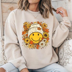 May include: Beige sweatshirt featuring a smiling face wearing a white knit hat with a pumpkin accent. The graphic is surrounded by yellow and orange flowers and fall leaves. The sweatshirt has long sleeves.