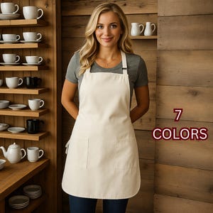 May include: A cream-colored apron with two front pockets and a neck strap. The apron is worn by a person in front of a wooden background. The text "7 COLORS" is displayed in a maroon font.