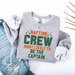 May include: A heather gray sweatshirt with the text "RAFTING CREW MOST LIKELY TO BE THE CAPTAIN" in green and orange. The sweatshirt has a crew neck and long sleeves. A silver necklace is draped across the top. The sweatshirt is folded next to a pair of blue jeans.