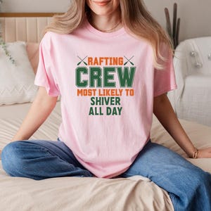 May include: A light pink t-shirt with the text "RAFTING CREW MOST LIKELY TO SHIVER ALL DAY" in green and orange. The shirt has a crew neck and short sleeves. The person is wearing blue jeans.