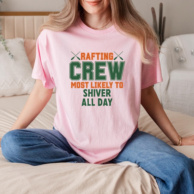 May include: A light pink t-shirt with the text "RAFTING CREW MOST LIKELY TO SHIVER ALL DAY" in green and orange. The shirt has a crew neck and short sleeves. The person is wearing blue jeans.