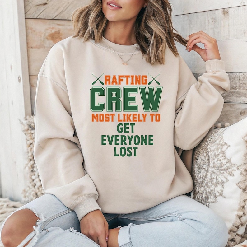May include: A beige sweatshirt with the text "RAFTING CREW MOST LIKELY TO GET EVERYONE LOST" in green and orange. The word "RAFTING" is above "CREW" with crossed paddles. The sweatshirt has long sleeves and a crew neck.