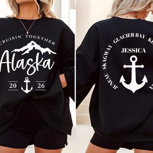 Puede incluir: Sudadera negra con texto gr&aacute;fico blanco y dise&ntilde;o de ancla. La parte delantera dice "Cruisin' Together Alaska 2026" con un gr&aacute;fico de monta&ntilde;a. La parte trasera presenta un dise&ntilde;o circular con "Jessica" y un ancla, rodeado de nombres de ciudades.