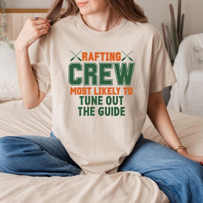 May include: Beige t-shirt with the text "RAFTING CREW MOST LIKELY TO TUNE OUT THE GUIDE" in green and orange. The design includes crossed paddles above the word "CREW". The shirt is a casual, crew neck style.