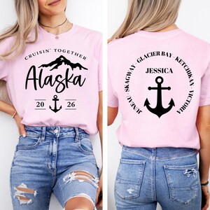 Puede incluir: Camisetas rosas con texto y gr&aacute;ficos negros. La parte delantera dice "Cruisin' Together Alaska 20 26" con un ancla. La parte trasera tiene un dise&ntilde;o circular con "Jessica" y un ancla, con nombres de lugares.