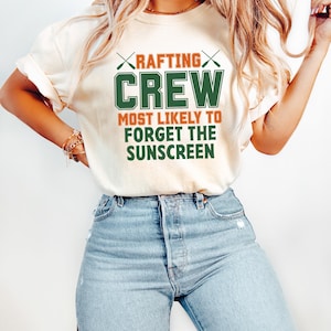 May include: Cream-colored t-shirt with the text "RAFTING CREW MOST LIKELY TO FORGET THE SUNSCREEN" in green and orange. The design includes crossed paddles above the word "RAFTING". The shirt is paired with blue jeans.