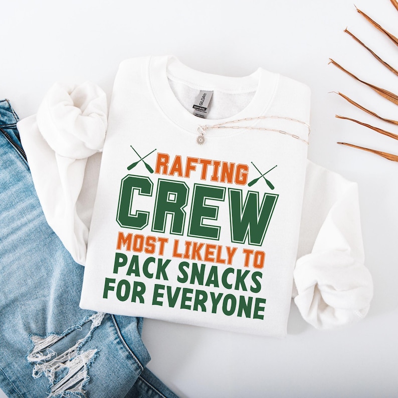 May include: White sweatshirt with the text "RAFTING CREW MOST LIKELY TO PACK SNACKS FOR EVERYONE" in orange and green. The design includes crossed paddles. A silver necklace with a pendant is visible.