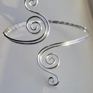 May include: A silver-toned arm cuff with a decorative, swirling design. The cuff features two prominent spiral accents and a delicate beaded band. The metal reflects light, creating a bright and elegant appearance. This piece is a stylish accessory.