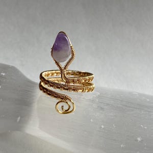 以下が含まれることがあります： 金色のワイヤーラップリング、スパイラルデザイン。 リングは、上部に涙の形をした紫色のアメジスト宝石が特徴です。 リングは白く反射する表面に表示されています。