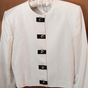 May include: A white jacket with a round neckline and long sleeves. The jacket has five dark navy blue rectangular accents with gold-coloured buttons. It is hanging on a white satin hanger.