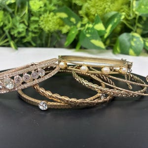 May include: A collection of gold-toned bracelets. One bracelet features clear gemstones, while another has small pearls. Other bracelets have textured or braided designs. The bracelets are displayed on a black surface.