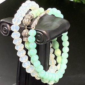 May include: Three beaded bracelets in shades of white and green are displayed on a black pedestal. The bracelets feature round and faceted beads. The bracelets are stacked together, showcasing the different textures and colors of the beads.