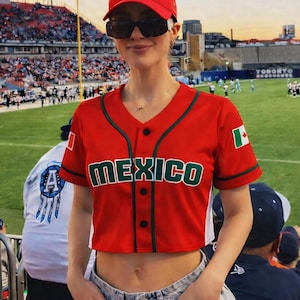 May include: A red baseball jersey with the word "MEXICO" in green lettering. The jersey has a green trim and a Mexican flag patch on the sleeve. The person is wearing a red baseball cap and sunglasses.