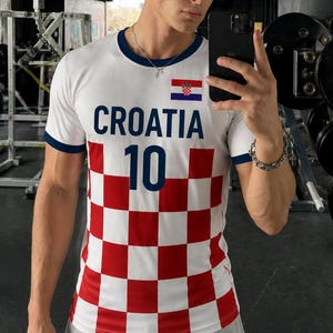 K&ouml;nnte beinhalten: Wei&szlig;es T-Shirt mit marineblauen Verzierungen und dem Wort "CROATIA" in marineblauen Gro&szlig;buchstaben. Das Shirt zeigt die kroatische Flagge und die Nummer "10" in Marineblau. Der untere Teil des Shirts hat ein rot-wei&szlig;es Karomuster.