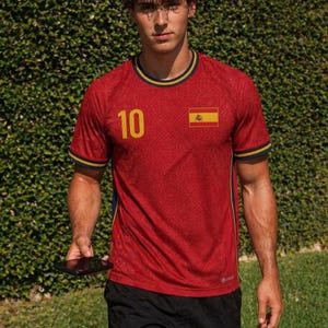 Op de afbeelding: Rood voetbalshirt met het nummer 10 in geel en de Spaanse vlag op de rechterborst. Het shirt heeft marineblauwe en gele afwerking op de kraag en mouwen. De persoon draagt een zwarte korte broek.