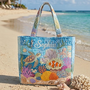 May include: A blue tote bag with an underwater design, including a sea turtle, clownfish, and coral. The bag has a name embroidered on it and is on a sandy beach with seashells.