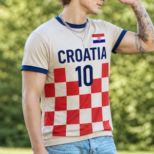 K&ouml;nnte beinhalten: Beiges T-Shirt mit marineblauen Verzierungen, mit dem Wort "CROATIA" in Marineblau, der Nummer "10" und einer kroatischen Flagge. Der untere Teil des Shirts hat ein rot-wei&szlig;es Schachbrettmuster.