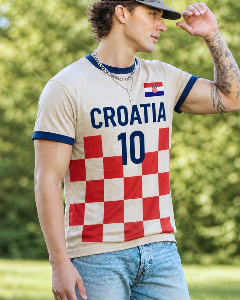 K&ouml;nnte beinhalten: Beiges T-Shirt mit marineblauen Verzierungen, mit dem Wort "CROATIA" in Marineblau, der Nummer "10" und einer kroatischen Flagge. Der untere Teil des Shirts hat ein rot-wei&szlig;es Schachbrettmuster.