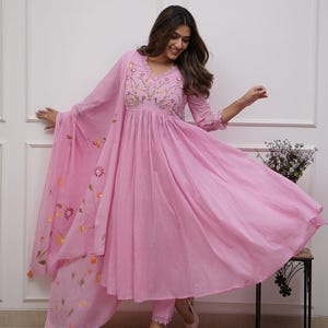Könnte beinhalten: Hellrosa Anarkali-Anzug mit floraler Stickerei auf dem Oberteil und Dupatta. Das langärmlige Kleid hat einen fließenden Rock und wird mit einer passenden Hose kombiniert. Der Dupatta hat florale Akzente. Das Outfit wird mit cremefarbenen Absätzen abgerundet.