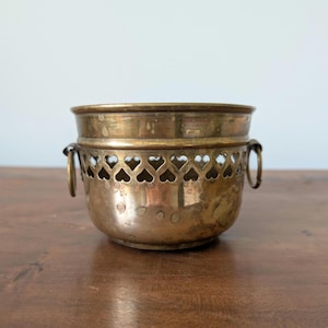 May include: A round brass bowl with a weathered patina, featuring a decorative band of cut-out heart shapes along the upper rim and two small circular ring handles attached to the sides. The metal surface shows signs of age and oxidation.