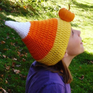 May include: A handmade crochet hat in the shape of a candy corn, featuring white, orange, and yellow sections. A small orange pumpkin sits atop the hat. The hat is worn by a person in a purple shirt, with a green and brown background.