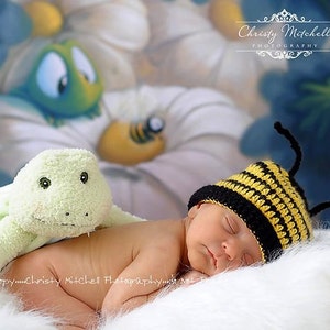 May include: A newborn baby wearing a yellow and black crocheted bee hat with antennas sleeps on a white fluffy blanket. A green plush turtle toy is beside the baby.