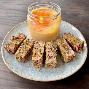 Puede incluir: Un plato gris claro contiene seis porciones rectangulares de comida, cada una cubierta de hierbas y pétalos de flores. Un tarro de cristal con una vela dentro está sobre el plato, emitiendo un brillo cálido. Las porciones son de color marrón rosado.