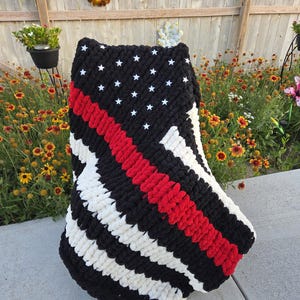 May include: A chunky knit blanket featuring a black field with white stars, a red stripe, and alternating black and white stripes. The blanket is displayed outdoors, draped over an object.