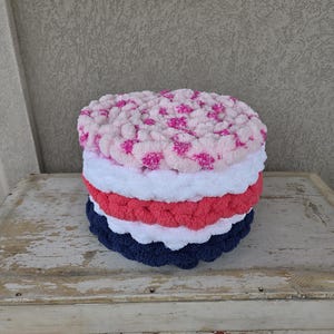 May include: A stack of handmade, round, plush items in various colors. The top layer is light pink with darker pink accents. Below it are layers of white, coral, and navy blue. The items are sitting on a distressed, white wooden surface.