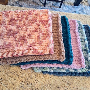 May include: A stack of six handmade, chunky knit blankets in various colors. The blankets include shades of pink, brown, teal, light pink, green, and blue. Each blanket has a textured, looped appearance.