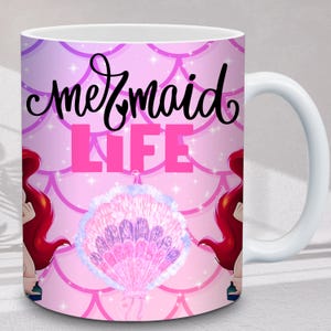 Puede incluir: Taza de cer&aacute;mica blanca con tem&aacute;tica de sirenas. La taza presenta las palabras "mermaid LIFE" en negro y rosa, junto con una ilustraci&oacute;n de concha. El fondo es rosa con un patr&oacute;n de escamas y detalles de estrellas. Ilustraciones de sirenas pelirrojas a cada lado.