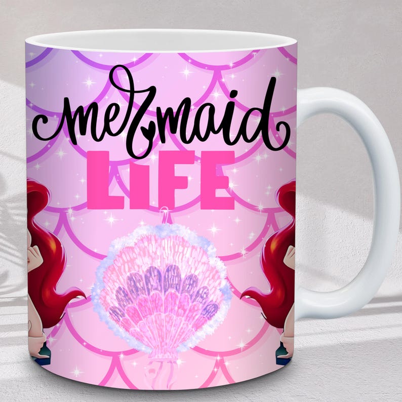 Puede incluir: Taza de cer&aacute;mica blanca con tem&aacute;tica de sirenas. La taza presenta las palabras "mermaid LIFE" en negro y rosa, junto con una ilustraci&oacute;n de concha. El fondo es rosa con un patr&oacute;n de escamas y detalles de estrellas. Ilustraciones de sirenas pelirrojas a cada lado.
