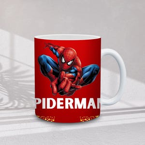 May include: White ceramic mug featuring a vibrant image of Spider-Man in a dynamic pose against a red background. The word "SPIDERMAN" is printed in white. The mug has a white handle and a gold star pattern at the bottom.