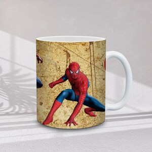 May include: White ceramic mug featuring a Spiderman design. The mug has a beige background with a distressed texture and a Spiderman graphic in red and blue. The handle is white.