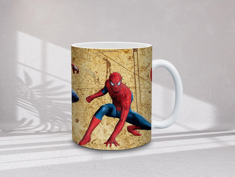 May include: White ceramic mug featuring a Spiderman design. The mug has a beige background with a distressed texture and a Spiderman graphic in red and blue. The handle is white.