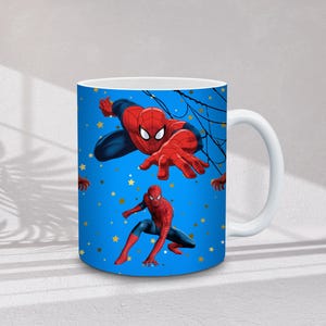 May include: White ceramic mug featuring Spider-Man in action poses against a blue background with gold stars. The superhero is depicted in his classic red and blue suit, with web details.