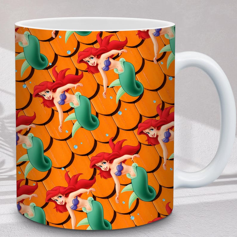 Puede incluir: Taza de cer&aacute;mica blanca con un patr&oacute;n repetido de una sirena de dibujos animados con cabello rojo, un top de concha azul y una cola verde. El fondo es un patr&oacute;n de escamas naranja. La taza tiene un asa blanca.