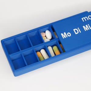 May include: A blue pill organizer with multiple compartments, some containing pills and capsules of various colors. The lid is open, displaying the days of the week and time designations in white text. The organizer is on a white surface.