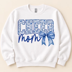 May include: A white crewneck sweatshirt featuring the text CHEER in a blue floral pattern and the word mom in blue script. A large blue bow graphic is positioned to the right of the text. The garment has long sleeves and a ribbed neckline.