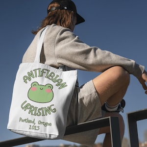 May include: White tote bag featuring the words "ANTIFIBIAN UPRISING" and a cartoon frog design. The bag also includes the text "Portland, Oregon 2015". The bag has white straps and is made of a canvas-like material.