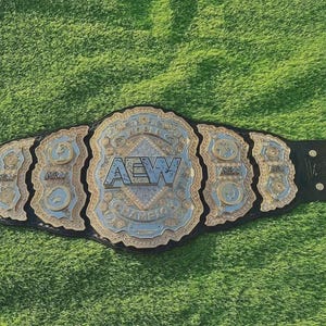 May include: A black leather championship belt featuring a large central silver and gold plate with the letters AEW and the word CHAMPION engraved. The belt includes multiple smaller side plates with intricate detailing and gold-toned metal studs.
