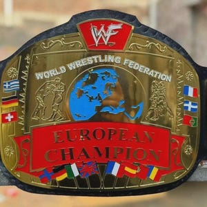May include: A gold-plated World Wrestling Federation European Champion wrestling belt featuring a central blue globe graphic, red banners with the text EUROPEAN CHAMPION, and various international flags on a black leather strap with side plates.