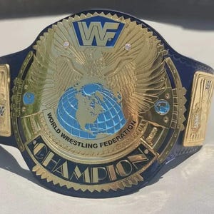 May include: A championship wrestling belt featuring a dark blue leather strap and a large gold-plated center plate. The plate displays a central blue globe, an eagle with spread wings, the WWF logo, and the text World Wrestling Federation Champion.