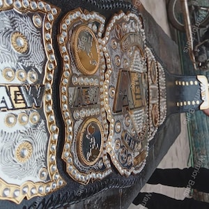 May include: A black leather championship belt featuring large, ornate gold and silver metal plates. The central plate displays the letters AEW in bold, raised lettering, surrounded by intricate engravings, circular gold accents, and clear crystals.